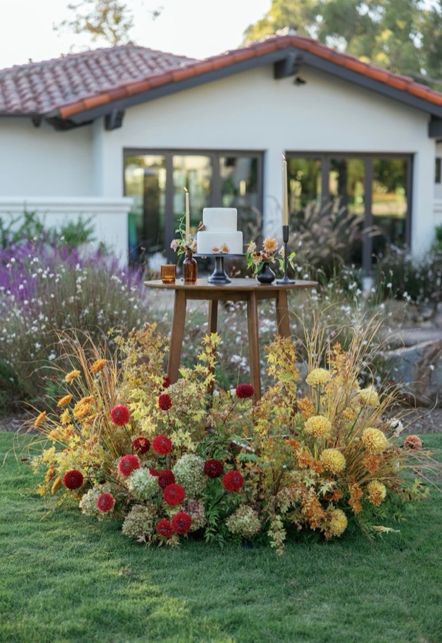 Autumn Romance wedding cake display — white tiered cake surrounded by a lush ground-level floral arrangement of dahlias and grasses in rust and gold — Lux Events by Michelle