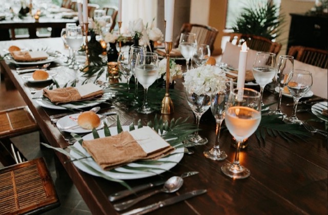 Coastal Celebrations tablescape — long farmhouse table with palm frond runner, gold candelabras, and white hydrangeas in candlelit setting — Lux Events by Michelle