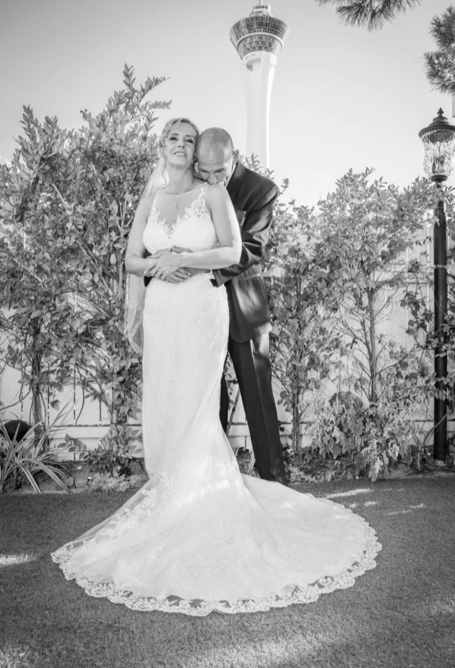 Destination wedding portrait in black and white — groom embraces bride in cathedral lace gown, Stratosphere Tower visible in background — Lux Events by Michelle